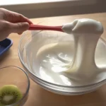 person lifting thick stretchy yogurt with a red spatula from a glass bowl on a wooden table