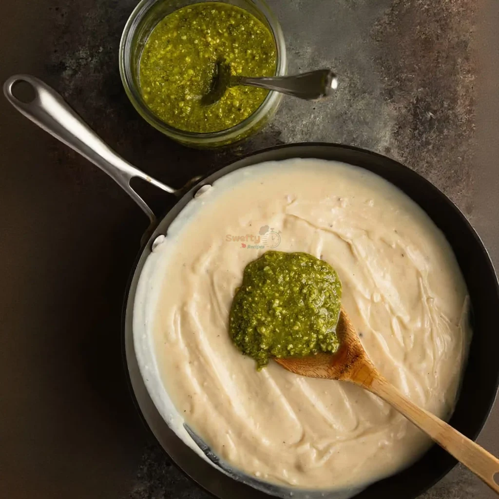skillet filled with creamy Pesto Alfredo sauce topped with a spoonful of fresh green pesto and a wooden spoon beside a jar of pesto